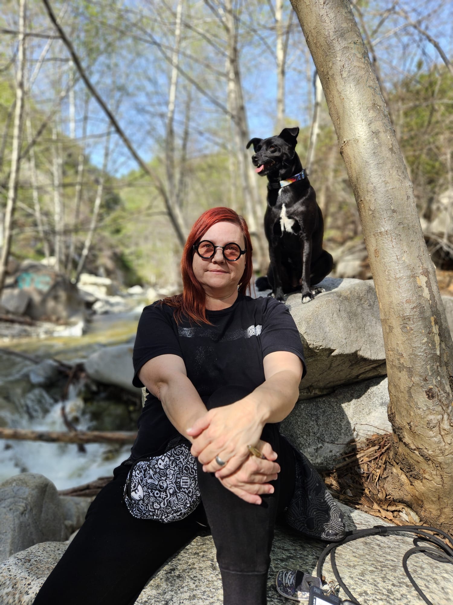 Paula Kelley seated on rocks beside a stream with her black dog